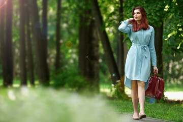 Obraz na płótnie Canvas a young beautiful girl of Caucasian race in a light blue dress and a red bag in her hands is walking along a path in a green forest. a woman walks in nature and adjusts her red hair with her hand