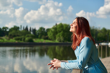 Naklejka na ściany i meble a beautiful young girl stands in nature and looks at the sun with her eyes closed. a girl with red hair enjoys summer and warmth