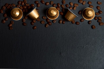 Caffeine, hot drinks and objects concept. Close up golden capsules or pods for coffee mashine with some roasted grains on black graphite background. Top view with space for text. Selective Focus.