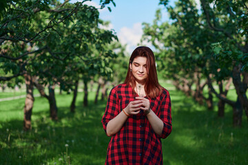 Fototapeta premium a young girl in a black and red checked shirt with red hair stands on a natural green background in a Park and holds a white dandelion in her hands, looks at it and smiles