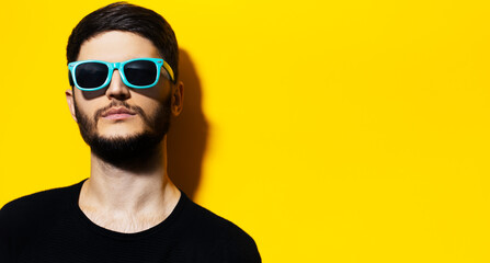 Studio portrait of young serious confident guy wearing cyan sunglasses on background of yellow color with copy space.