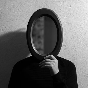 Fototapeta Black and white studio portrait of young man holding oval mirror on face. Background of textured wall.