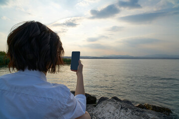 Woman on vacation on lake garda photographs the beauty of the sunset with her smartphone. The water is clear and calm, the sky is full of clouds, the sun is reflected on the water.