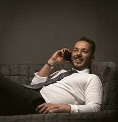 Fototapeta premium Businessman talks over mobile phone lying on sofa. Smiling man at couch talking smartphone on grey back. Close up shot. Toned image.