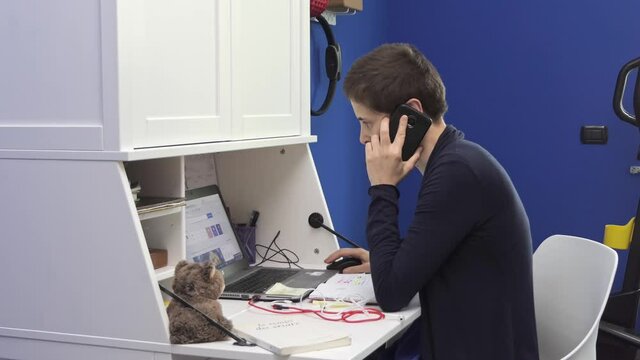 Young woman answers the smartphone while she is in smart working at home in the room, sitting at the desk in front of the laptop