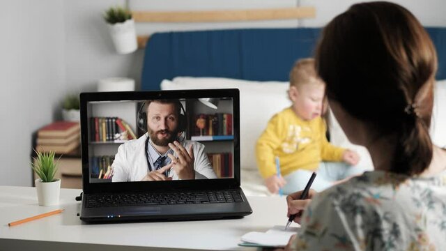 Doctor online consultation video link. Woman looks at laptop screen and talks to Internet streaming connection with man in white coat. In background is little kid playing. Close-up