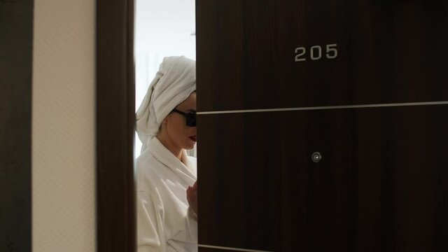 a girl in red earrings and sunglasses with a towel on her head and a white coat opens a 205 room in a hotel, looks at the camera and smiles
