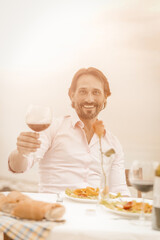 Smiling handsome holds glass of red wine looking at camera while sitting outdoor at table on sea background. Celebratory toast concept. Vacation concept. Toned image.
