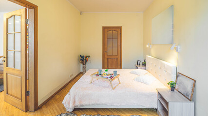 Modern light interior of bedroom in apartment. Side view of the  bed and bedsides. Breakfast on a tray. Wooden doors.