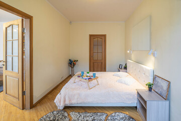 Modern light interior of bedroom in apartment. Side view of the  bed and bedsides. Breakfast on a tray. Wooden doors.