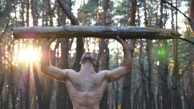 Muscular man lifting hard weight over head training hands. Strong guy with naked torso working out in forest. Athlete exercising at beautiful nature environment. Sport and active lifestyle. Dolly shot
