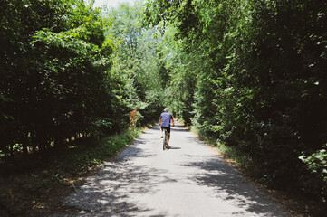 Rear view of active senior man riding bicycle along forest road on summer day - Senior athletic man cyclist riding bike on the trail in sunny forest - Healthy lifestyle and travel concept - Copy space
