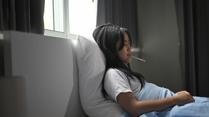 Fototapeta premium A little sick girl covered in a blanket is lying on the bed and measuring the temperature.