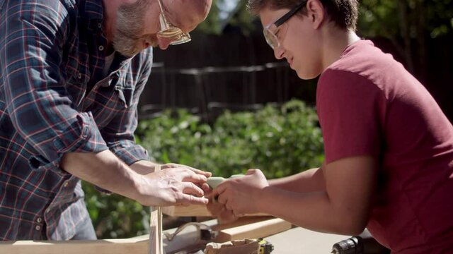 A father helps his son drive a screw in with a hand drill while working on a DIY carpentry project