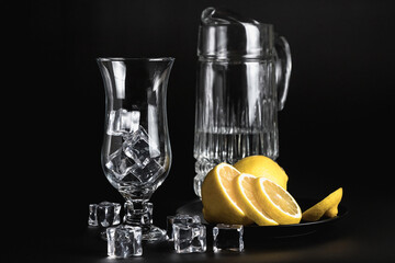 Sliced lemon on a black plate. A decanter and a tall glass of water and ice. Still life of glass...
