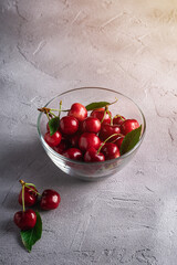 Fresh ripe cherry fruits with green leaves in glass bowl, summer vitamin berries on grey stone background, angle view