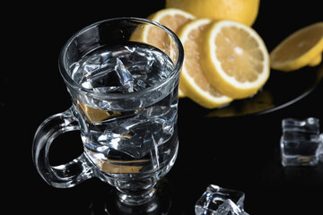 Sliced lemon on a black plate. A decanter and a tall glass of water and ice. Still life of glass objects on a black background. Cold lemonade. Yellow on black.