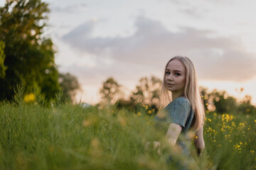 Fototapeta na wymiar Young happy blonde girl staying at a green wheat field in the evening against the background of the sun shines. Concept of freedom. Summer time joy. Beautiful young woman walking on field.