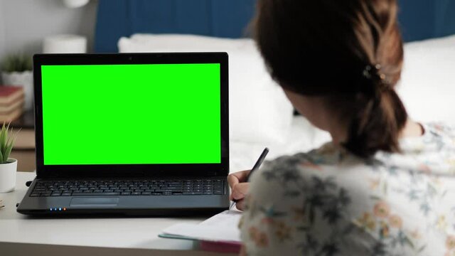 Woman sits at desk in bedroom, she looks at laptop green screen and talks to someone over internet video communications, sometimes taking notes in notebook. Close-up
