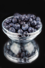 A clear glass bowl of blueberries on a mirror with reflection.  Black background.