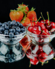 Three glass bowl filled with strawberries, cherries, and blueberries on a mirrow with reflection.  Black background.