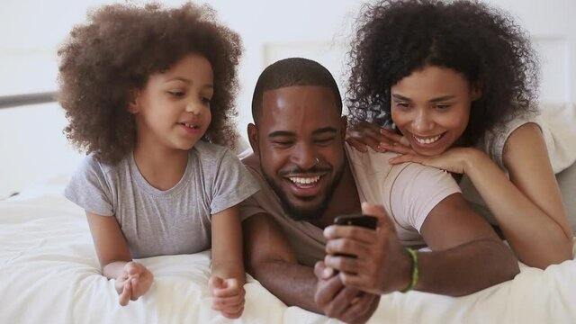 Happy african family with small cute kid daughter having fun hold phone using funny app looking at cellphone screen, cheerful parents and little child girl take selfie laugh lying on bed in morning