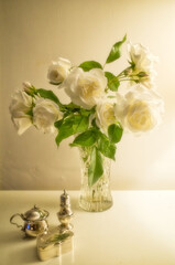 Bouquet of white roses in crystal glass vase with silver trinkets in the foreground. Soft focus.