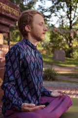 
Buddhist man in a purple shirt meditates on the background of a Buddhist statue
