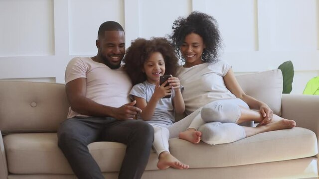 Happy african family with small cute kid daughter sit on sofa having fun with smart phone, little child girl holding cellphone using funny mobile app laughing with black parents take selfie at home