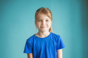 Close-up studio shot of a lovely blond little girl in a blue t-shirt posing against a blue background. Looks at the camera and smiles.