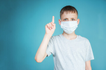 Portrait of a teenager in a gray t-shirt. Photo of a young boy on a blue background. He is in a medical mask, his index finger is raised up. The concept of virus, caution, safety. Copy space