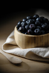 Fresh blueberries with small drops of water on the surface, in a bamboo bowl on a wooden table, with a tablecloth below and a wooden spoon.
