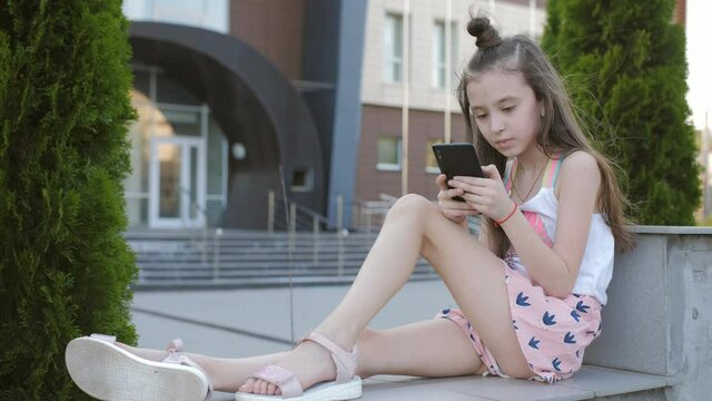A little elementary school girl sits on a parapet in the background of the school's campus building and uses a smartphone.