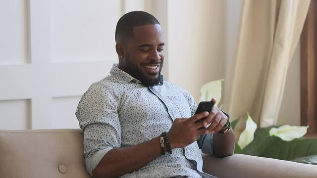 Smiling young black man holding phone using mobile apps for work education shopping online sit on sofa, happy african american guy texting sms message in social media play game on smartphone at home