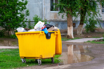 Trash bin, garbage container full of waste. Yellow trash can near the apartment building. Environmental pollution concept, bad ecology.