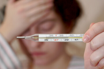  A young woman holds on to her forehead and shows a thermometer with fever.