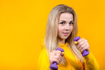 Portrait of beautiful young blonde woman in casual yellow sporty clothes doing exercises with small 0.5 kg purple dumbbells.