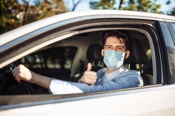 Young taxi driver shows thumb up sign sitting in the car and wearing protective sterile medical mask, works hard during coronavirus outbreak. Social distance, virus spread prevention, treat concept.