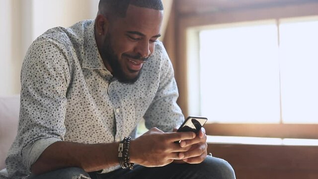 Young african american man holding smartphone device texting sms message sitting at home office, smiling black guy using apps playing mobile game chatting in social media surfing web on phone indoors