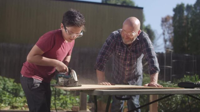 A teenage boy makes a cut with a circular saw as his father looks on in the backyard