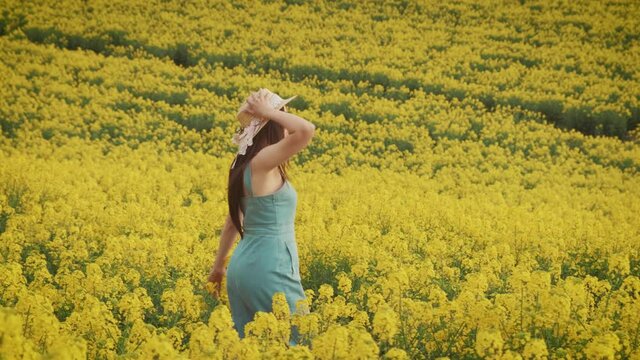 Happy young beautiful woman walks on yellow flowering field rapeseed, blooming summer flowers meadow. Brunette girl long hair enjoy recreation, spring nature, blue sky. Casual straw hat denim dress 
