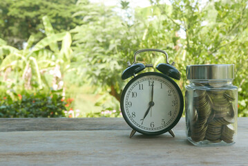 A vintage alarm clock and a jar full of gold coins on a wooden table on a bright sunny day with a nature background.