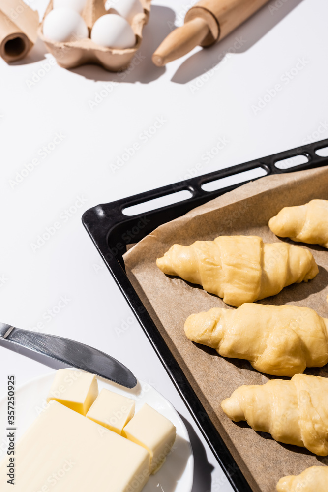 Wall mural selective focus of raw croissants on baking tray near rolling pin, parchment paper, butter, eggs, knife on white background