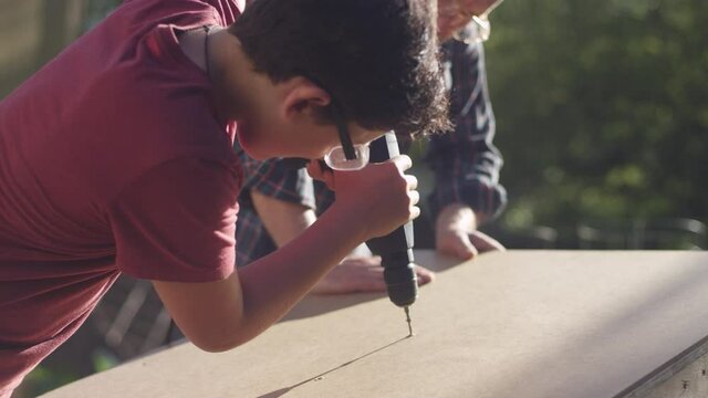 A father watches his teenage son driving a screw into a piece of wood with a power drill