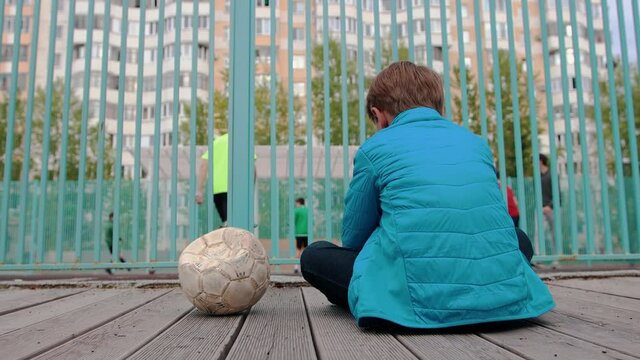 A little boy sitting on the bench near the deflated ball and watching other kids playing football