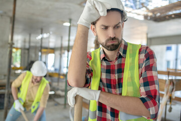 Tired building worker in yellow vest and gloves touching his helmet, sighing, his coworker working behing him