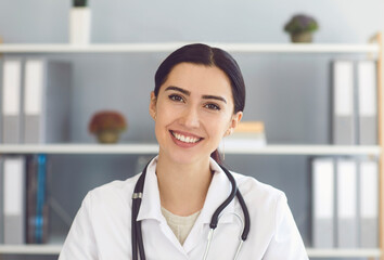 Portrait of beautiful young female doctor looking at camera in medical office, having online consultation with patient