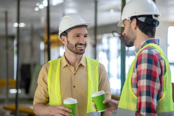 Building workers in yellow vests and helmets having coffee break, talking, smiling