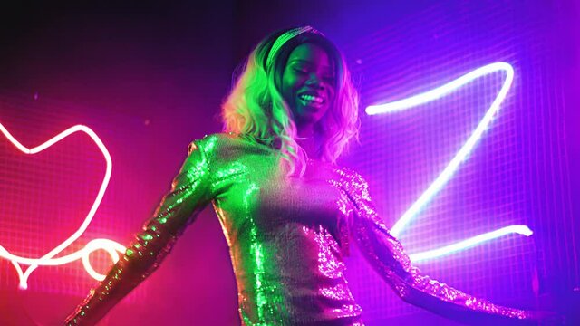 Happy African American girl is dancing in the neon lights of a night club or party at home in a silver sequin dress with neon figure lights background. Neon concept. Prores 422.