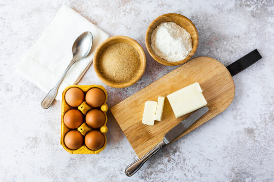 Napkin, spoon, bowls of flour and brown sugar, chicken eggs, cutting board, butter and table knife